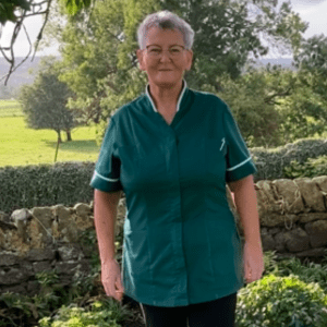 Gail Smart smiling in her green Herriot Hospice Homecare uniform in front of a dry stone wall and rolling hills of the dales.